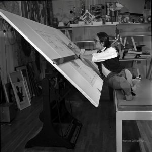 A black and white photo from 1966 of the artist François Dallegret sitting with his legs up sideways on a bench at his large drawing table, working on designs.