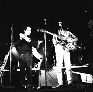 Black and white photograph of two members of the band Mothers of Invention in concert at The New Penelope, one plays a tambourine, the other man is Frank Zappa with a moustache and goatee and long black hair, playing electric guitar.