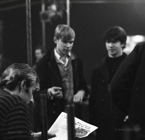 Photographie en noir et blanc de trois jeunes hommes, deux debout et un assis, dans le New Penelope. Au premier plan, l'un d'entre eux est assis sur un banc et lit un journal, à l'arrière-plan, les deux autres sont debout et le regardent. Le titre de l'article lu est 