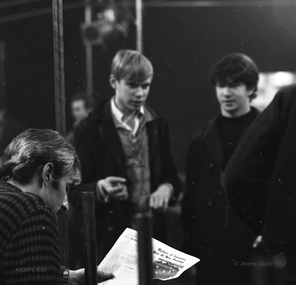 Black and white photograph of three young men, two standing and one sitting, in The New Penelope, in the foreground one of them is sitting on a bench reading a newspaper, in the background the two others are standing while looking at him. The headline of the article being read reads Mothers Of Invention Now at New Penelope.