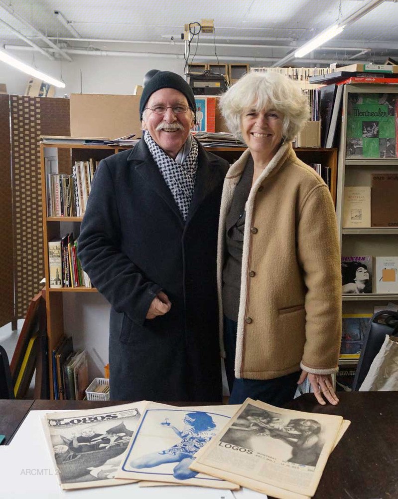 Photo of an older couple wearing winter coats and looking at the camera while standing in front of. bookshelves in a library with three copies of the newspaper Logos laid out on a table in front of them.