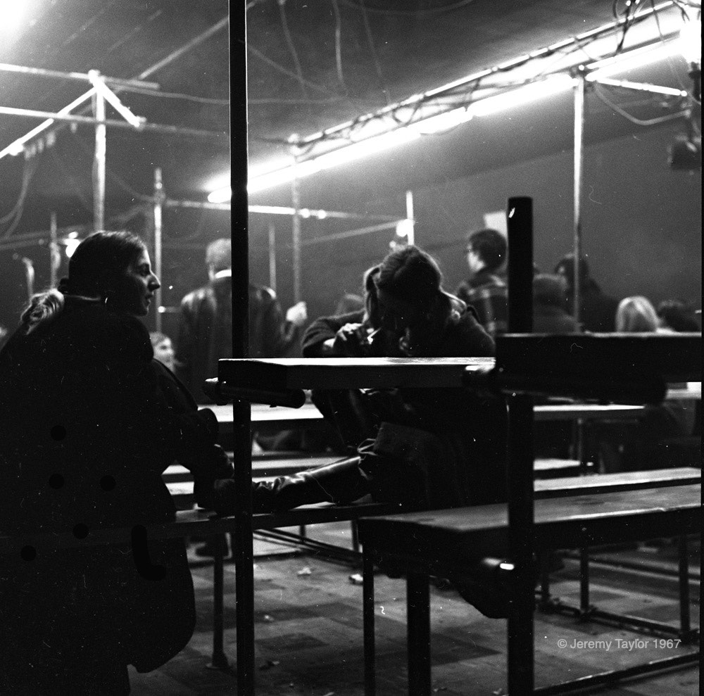 Black and white photograph of young people seated on benches at The New Penelope with two women in silhouette sitting in the foreground with their feet up on a bench. Bright lights are seen hanging off of scaffolding in the top of image.