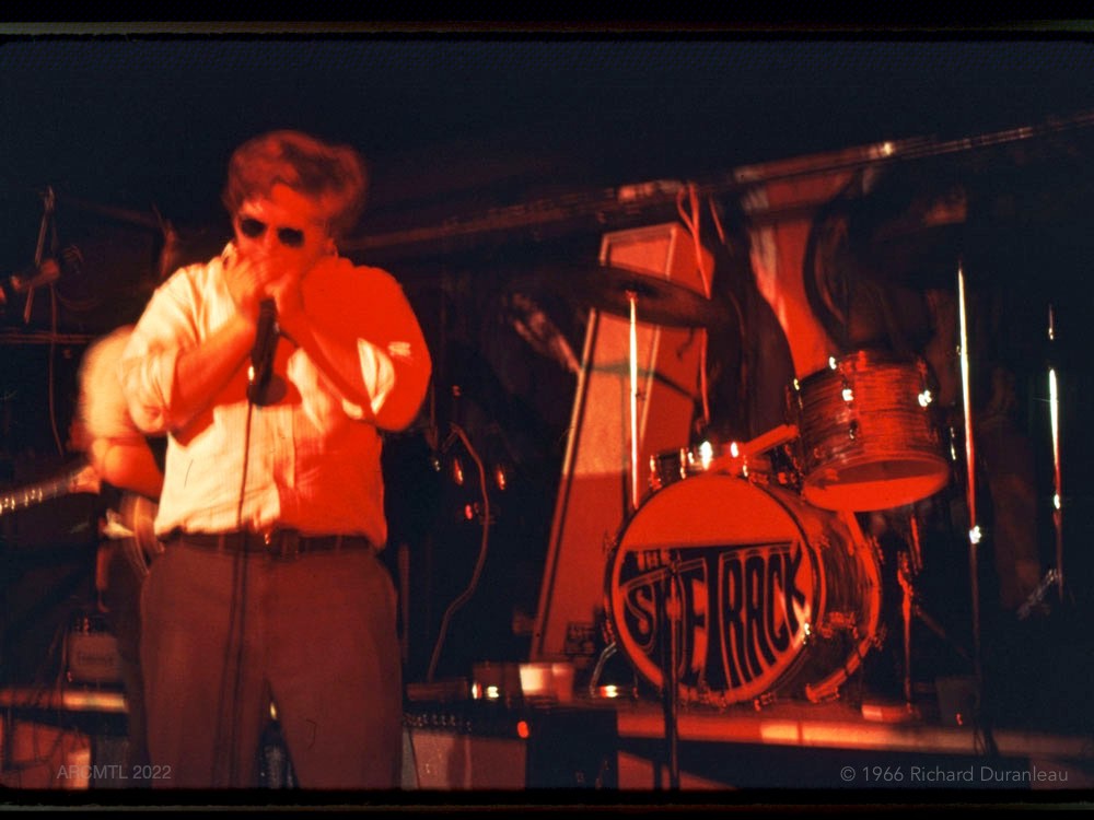 On a stage silhouetted in red, a man in white shirt, dark pants and sunglasses plays harmonica into a microphone. Beside him in the background, a drum kit with the name of the band on the drum, “The Sidetrack.”