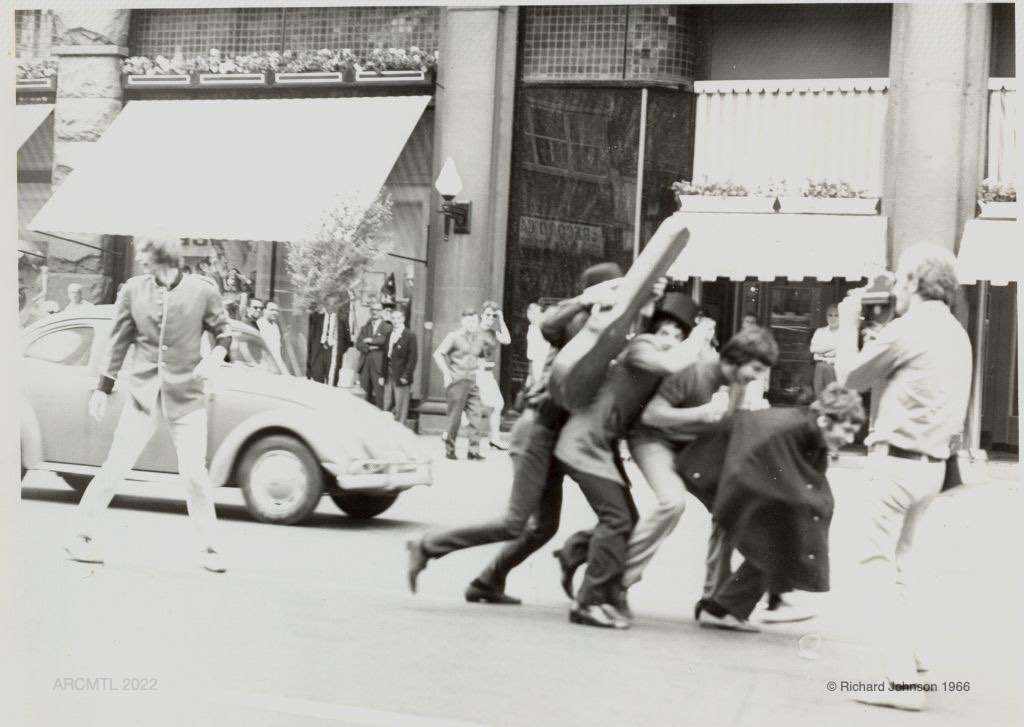 Black and white photograph of a group of young men smiling and hurrying down the middle of a commercial street wearing top hats, one of them holds a guitar case, a man is standing in their way filming them and in the background people on the sidewalk are watching the scene.