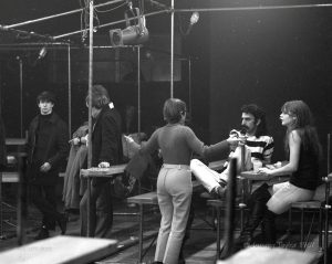 Black and white photograph of people standing and sitting while talking in The New Penelope. The musician Frank Zappa is sitting on a bench next to a young seated woman on the left side of the image, while listening to a woman with arms outstretched seen standing in front of him.