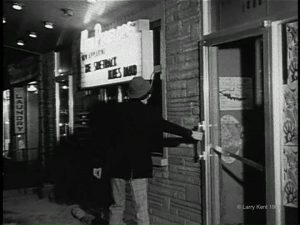 Capture d'écran en noir et blanc d'un homme avec un chapeau entrant dans un bâtiment avec une enseigne lumineuse à sa gauche annonçant le Sidetrack Blues Band au New Penelope.