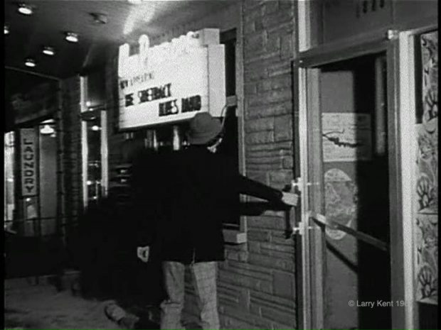 Black and white still image of a man with a hat entering a building with a lighted sign to his left advertising The Sidetrack Blues Band at The New Penelope.