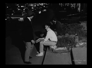 Black and white still image of a nightime view of Sherbrooke Street facing east with cars and headlights filling the background, in the foreground a man sits at the curb speaking with a second man wearing a hat standing up.