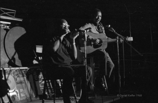 Black and white photograph of the performers Sonny Terry and Brownie McGhee sitting on chairs in the centre of a stage, one playing guitar and singing, one with an harmonica, behind microphones. The space is dimly lit.