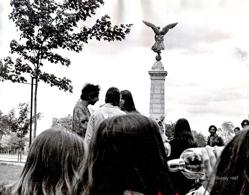 A black and white image of ten young people standing and sitting in a park around a statue in the centre of the image. The statue is of a stone winged angel atop a tall narrow stone pedestal.