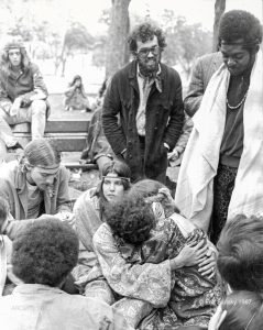 Photographie en noir et blanc de plusieurs jeunes gens assis sur des bancs dans un parc ainsi que sur le sol, avec un homme debout regardant un couple assis sur le sol s'étreignant au centre inférieur de l'image.