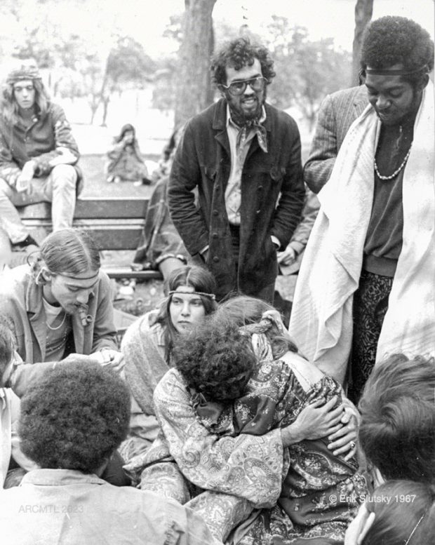 A black and white photograph of several young people sitting on benches in a park as well as on the ground, with a man standing and looking down at a couple seated on the ground hugging in the bottom centre of the image.