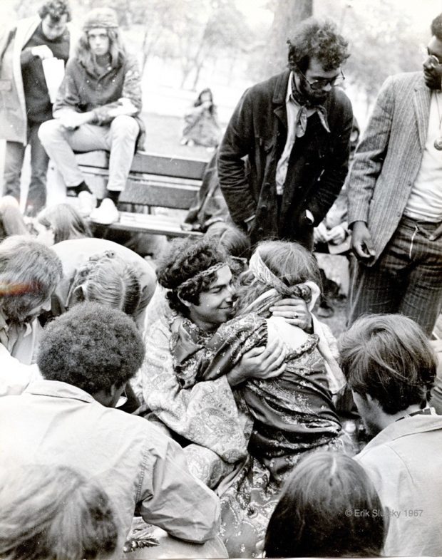 A black and white photograph of several young people sitting on benches in a park as well as on the ground, with a couple seated on the ground hugging in the centre of the image.