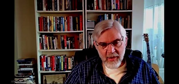 A screenshot of a video chat featuring Jim Mathewson seated in front of bookshelves and an acoustic guitar leaning against a window.