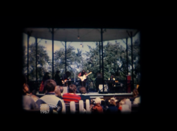 A screenshot from a video of the group Sidetrack performing outdoors in a gazebo in a Montreal park with trees visible behind them and a seated crowd of young people in front of them.