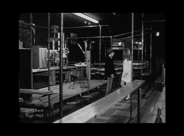 A still image taken from the film High showing a bench and scaffolding in front of the empty stage at The New Penelope with a man wearing a hat speaking with a young woman wearing a short white dress in the centre of the image.