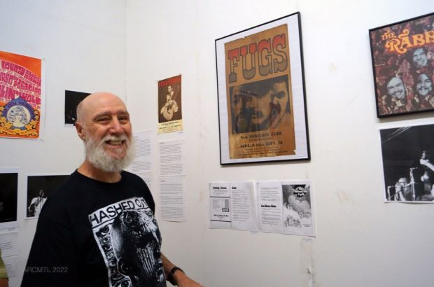 Allan Youster smiles at the camera white standing next to a framed poster advertising a concert at The New Penelope in an art gallery space