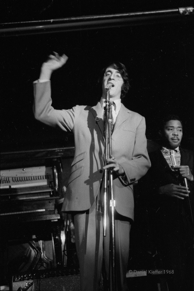 A tall man wearing glasses and a pale suit and tie stands at a microphone making an announcement on the stage at The New Penelope, with a musician visible behind him on one side and a piano on the other side.