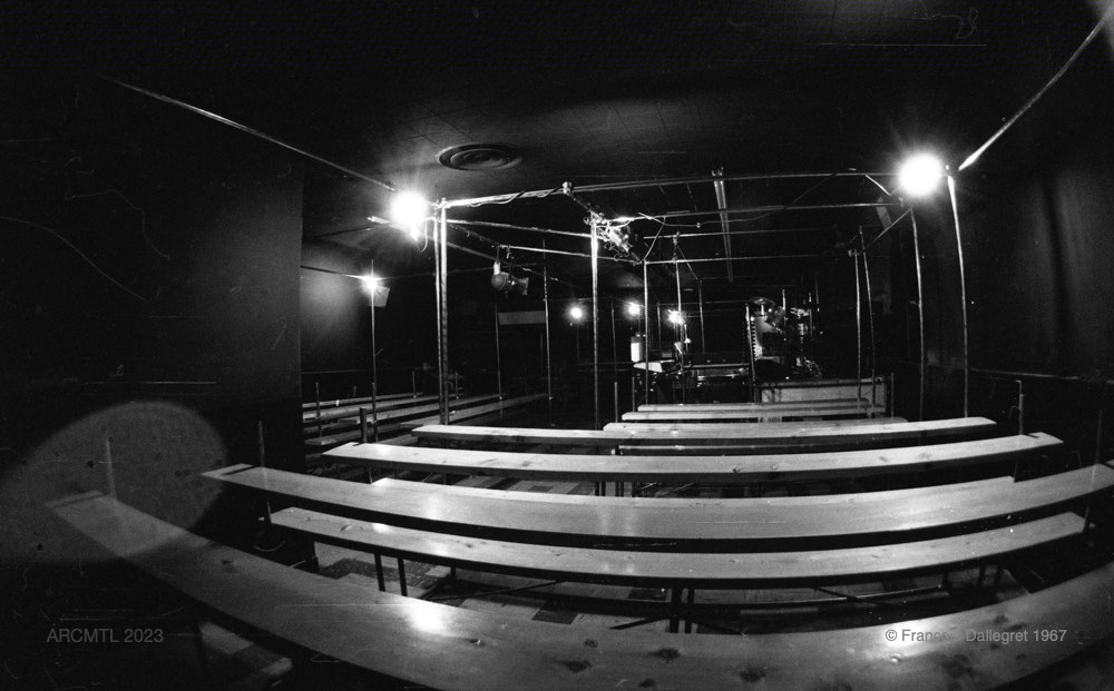 A black and white photograph of the interior of The New Penelope taken with a wide-angle lens showing a large empty space filled with rows of parallel wooden benches running from left to right.