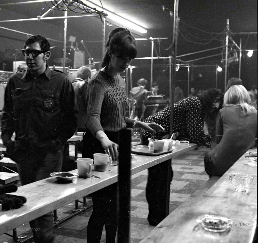 A black and white square photograph showing a brightly-lit space with several different groups of people socializing along long wooden benches. A young woman in the foreground is emptying an ashtray.