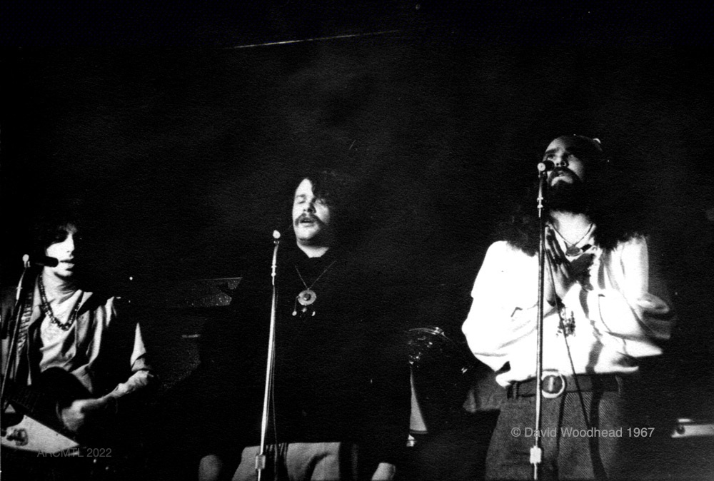 Black and white photograph featuring three members of The Fugs singing into microphones on the stage of The New Penelope, in the dark