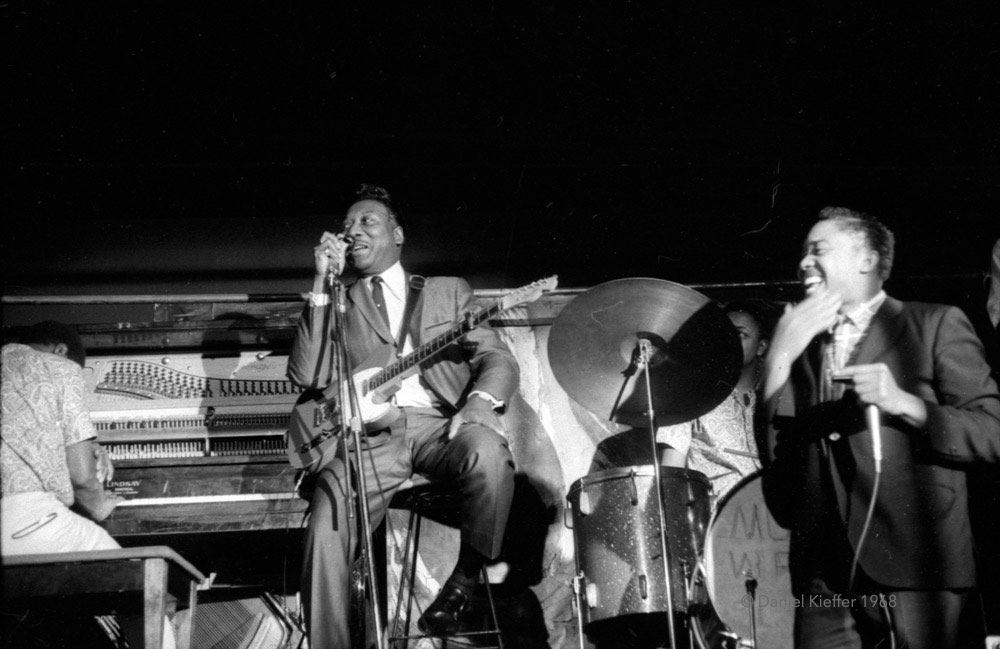 A black and white photograph showing blues singer Muddy Waters and his guitar seated in the centre of the stage wearing a suit and tie with his harmonica player to his left and his piano player to his right, performing at The New Penelope concert venue.