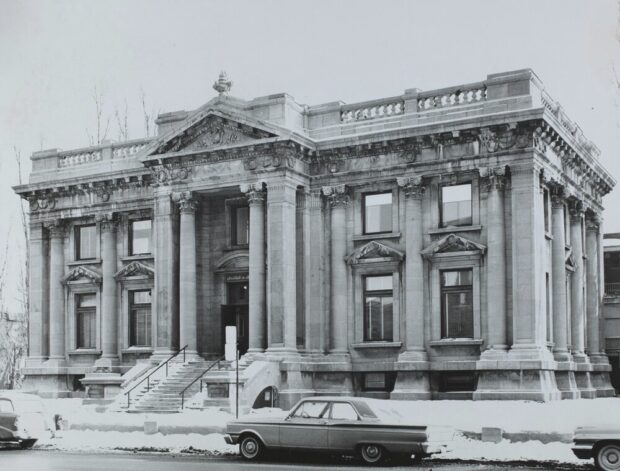 Black and white photograph of a winter urban landscape. The subject in the center of the image is a large beaux-arts inspired building made of large gray stone bricks. In the foreground, the street and a few parked cars are visible