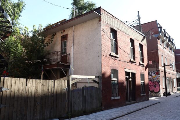 Color photograph of an alley with several back sides of buildings. In the center, the subject is a small duplex with an orange brick facade. Its door opens directly onto the alley, and it is bordered by a wooden fence.