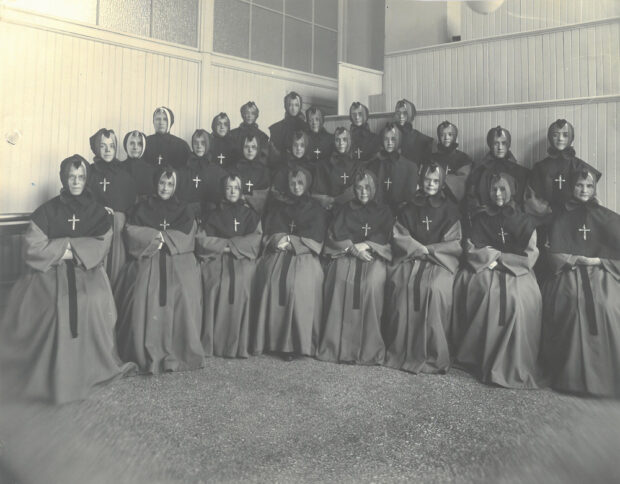 Black and white photograph of a group of 25 women in religious attire arranged in three tiers on a platform. They are in a room with wood panel walls and opaque glass windows at the top.