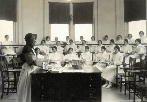 Black and white photograph of a round or octagonal room with windows on each wall and a high ceiling. The room is arranged in three-tiered stands. About twenty student nurses are seated at desk-chairs. They are wearing nursing uniforms. In the foreground, a woman in religious attire appears to be handling containers and pouring liquids.