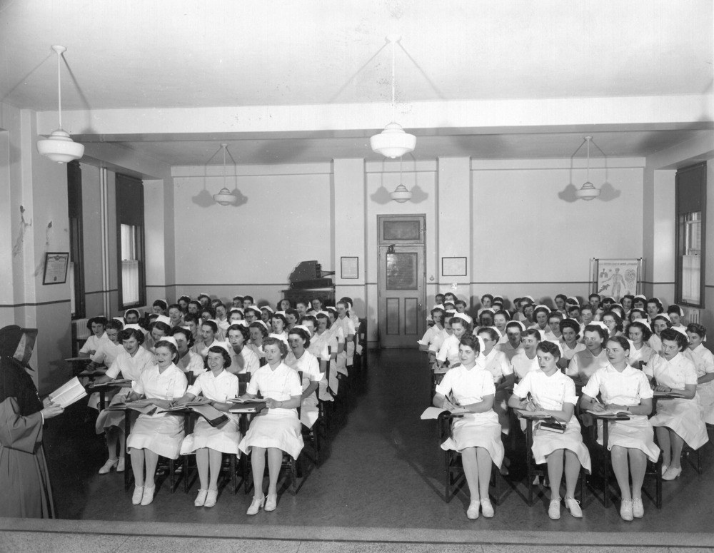 Black and white photograph of a large classroom. Windows line the side walls, the door is in the center of the back wall, and an anatomy chart is displayed at the back. In the foreground, a woman in religious attire holds an open book. More than sixty students in nursing uniforms are seated at desk-chairs, taking notes.