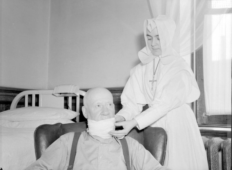 Black and white photograph of a woman in religious attire and a patient. She is standing behind her seated patient, changing his bandage located on his neck and lower face.