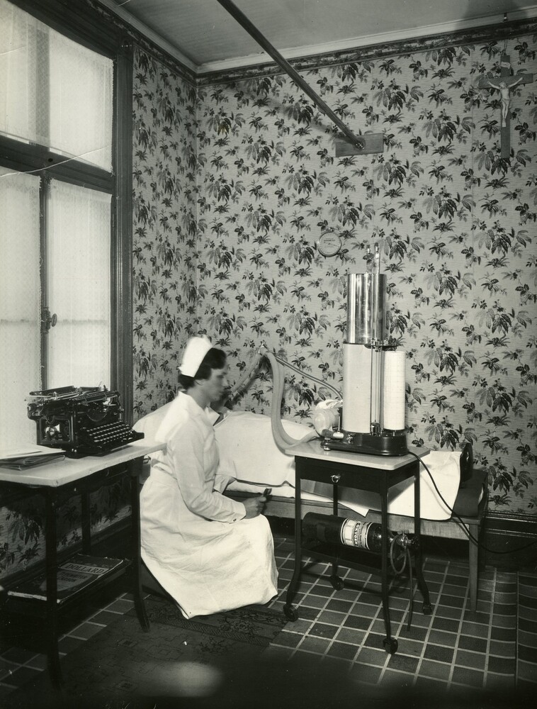 Black and white photograph of a small room with floral wallpaper. A bed, a chair, and a small table with a typewriter furnish the space. An unidentified patient is lying on the bed. A woman in a nurse's uniform appears to be maneuvering a tube connected to a machine towards the patient's face.