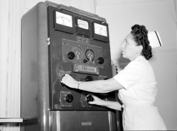 Black and white photograph of a woman in a nurse's uniform. She is adjusting rotary knobs on a large rectangular machine with several dials.