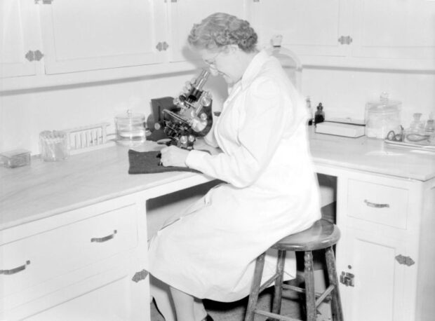 Black and white photograph taken in a room resembling a laboratory. A woman in a lab coat is seated on a bench, looking through a microscope..