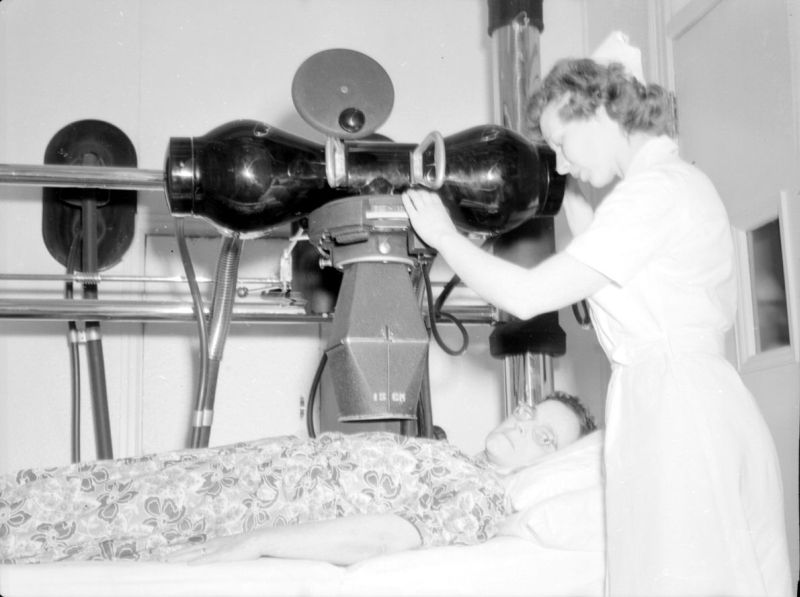 Black and white photograph depicting a small enclosed room painted white. A woman lying on her back on a hospital bed is receiving treatment from a machine aimed at her chest. This machine is operated by a second woman in a nurse's uniform. A large metal support is installed in the room, allowing the movement of the machine, which resembles a very large camera lens.