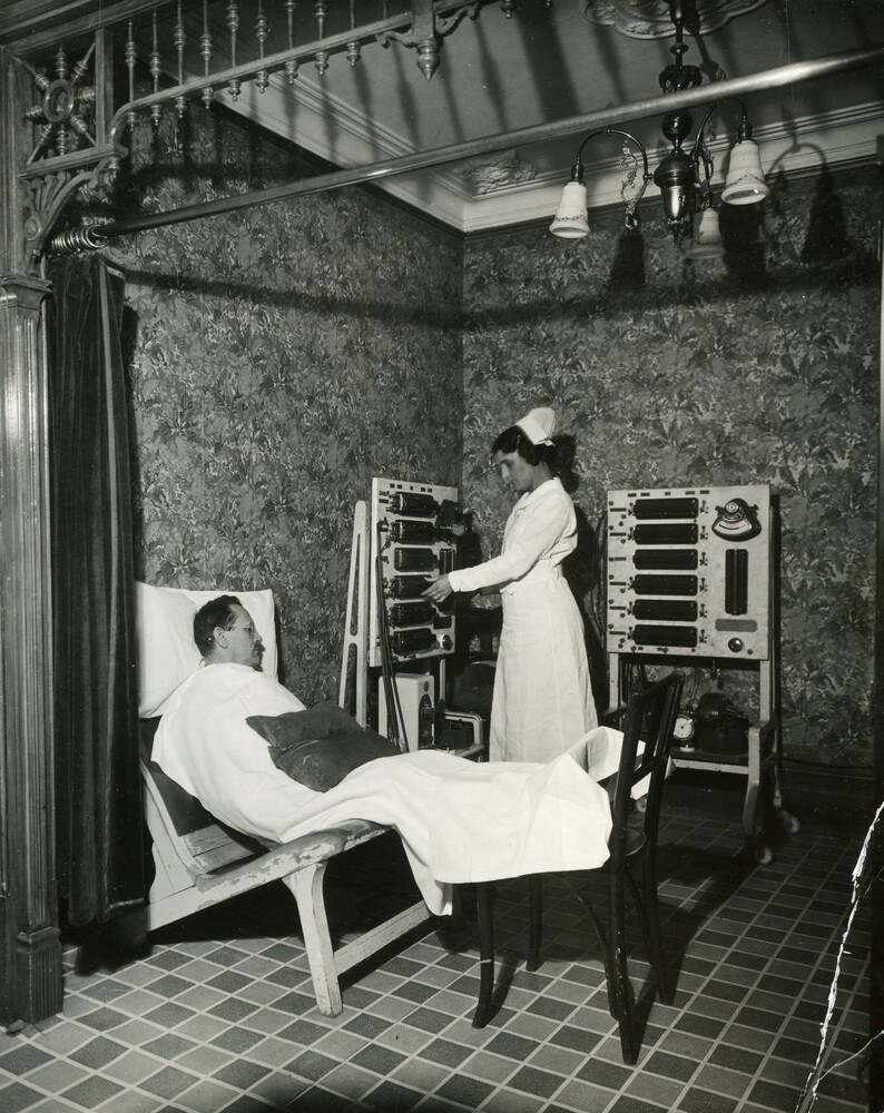 Black and white photograph of a room with lacquered wooden walls and tiled floors. The room is furnished with a hospital bed, cabinets, a wheeled table full of medical equipment, and several machines consisting of large glass and steel tanks.