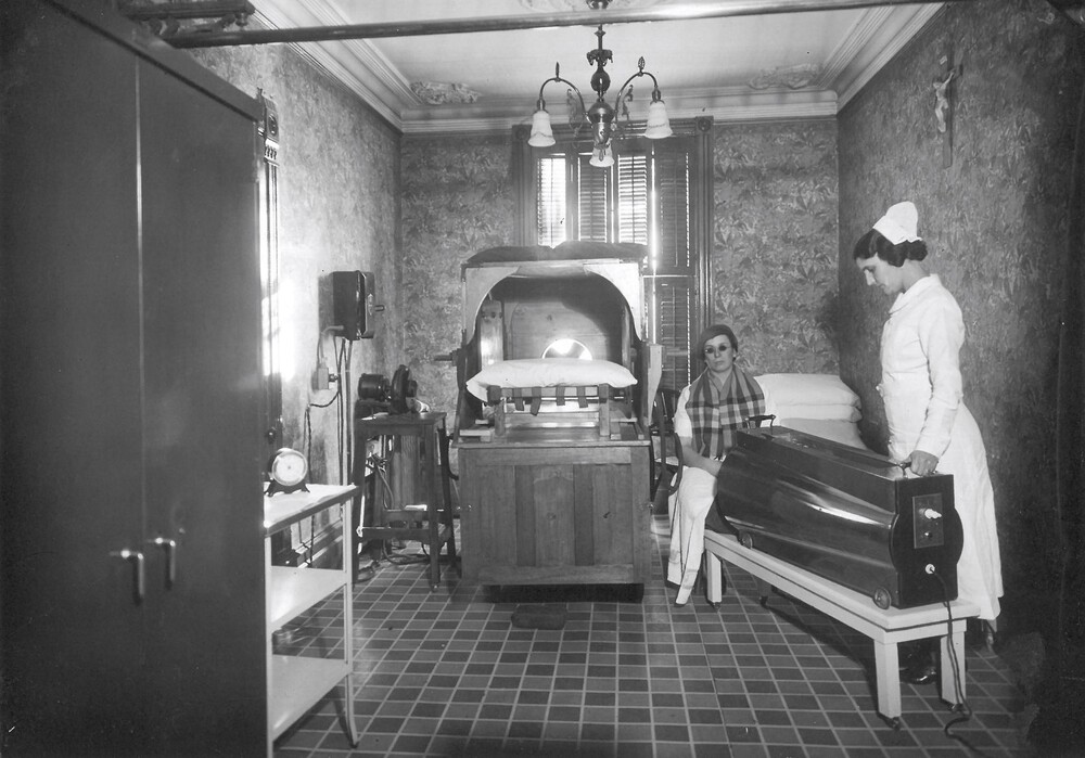 Black and white photograph depicting a small room with a window and closed shutters. There is a ceiling light and a crucifix on the wall. Some shelves hold small, unidentifiable devices. A sort of bed resembling a large wooden box with a pillow is in the center. A woman in a nurse's uniform points a machine resembling a dark cannon towards a person who appears to be a patient of the Institute.