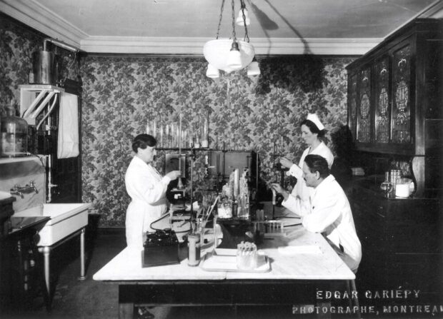 Black and white photograph of a windowless room with floral wallpaper. A lamp hangs from the ceiling, a large cabinet is on the right wall, and a large sink is on the left wall. In the center is a large table with an array of laboratory instruments. Test tubes, beakers, and other less recognizable instruments are handled by two women in nurse's uniforms and a man in a lab coat.