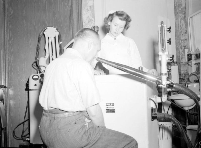 Black and white photograph featuring a woman standing in a nurse's uniform in the background and a man seated in the foreground. A box-shaped machine separates them. A tube emerges from the machine and is pointed at the seated man's face.