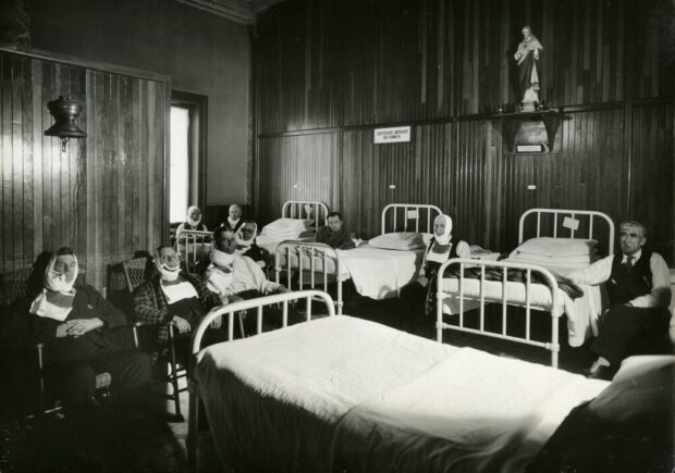 Black and white photograph depicting a room furnished with hospital beds and chairs. Nine men are seated, most with bandages around their necks or faces. A small statue of Jesus sits on a shelf, and a sign reading No smoking allowed is posted on the same wall.