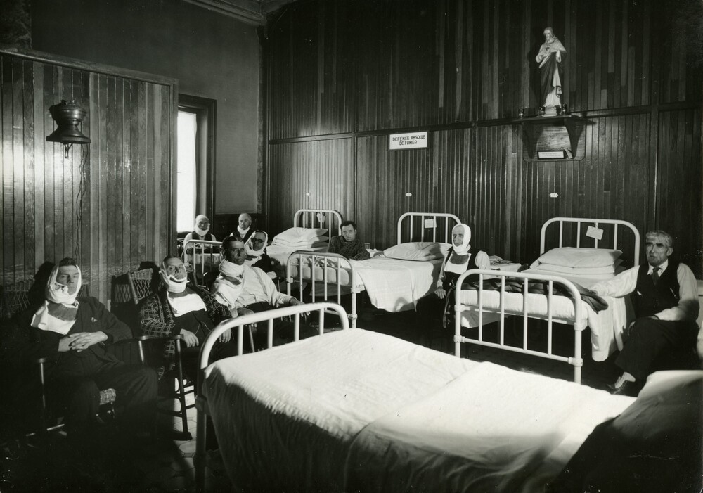 Black and white photograph depicting a room furnished with hospital beds and chairs. Nine men are seated, most with bandages around their necks or faces. A small statue of Jesus sits on a shelf, and a sign reading "No smoking allowed" is posted on the same wall.