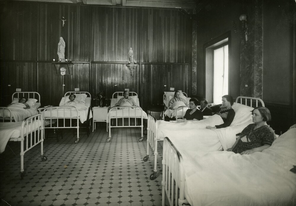 Black and white photograph depicting a room furnished with hospital beds and chairs. Two women are seated, and seven others are lying in the beds. Two small statues of Mary, the mother of Jesus, sit on shelves. A small crucifix is installed above them.