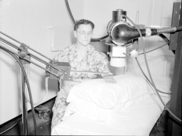 Black and white photograph depicting a seated woman placing her hand on a pillow on a hospital bed. Two machines are set up on stands around her. One is a metal rod held horizontally above her hand, and the other resembles a camera with its lens pointed over her hand.