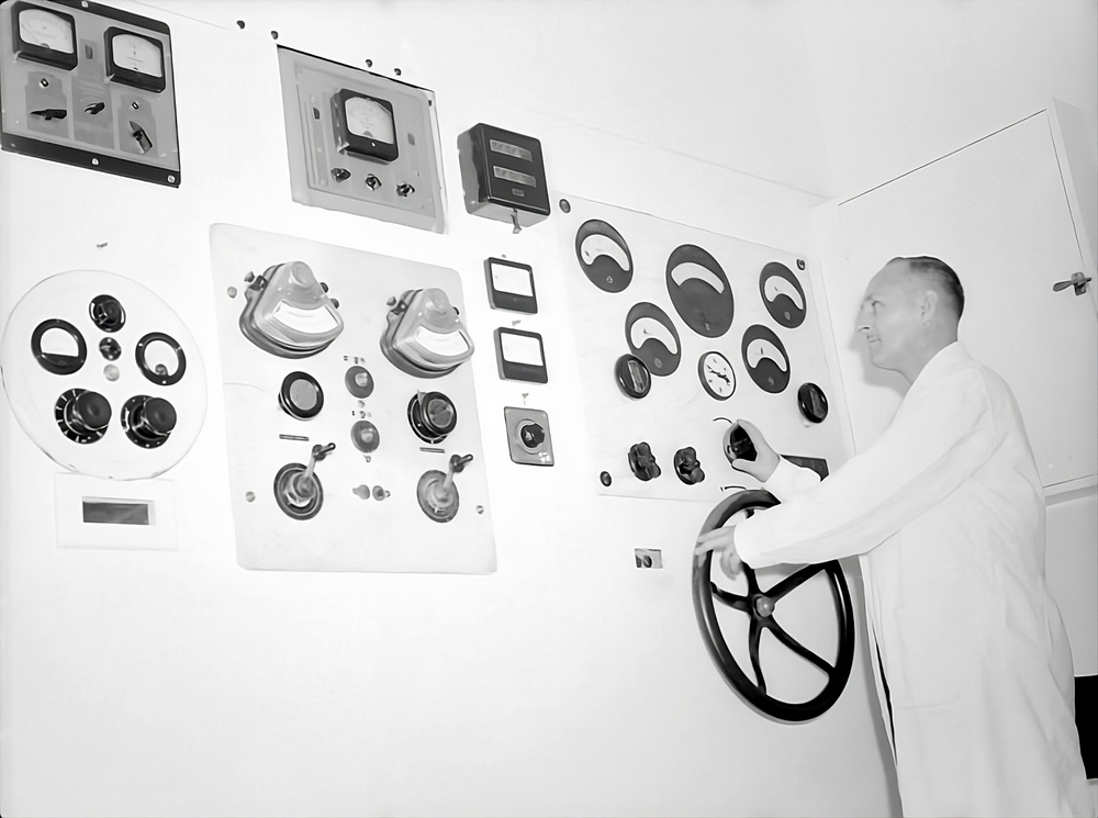 Black and white photograph of a man on the right side of the image using a large machine that occupies the rest of the space. The machine is a large white wall with various dials and levers.