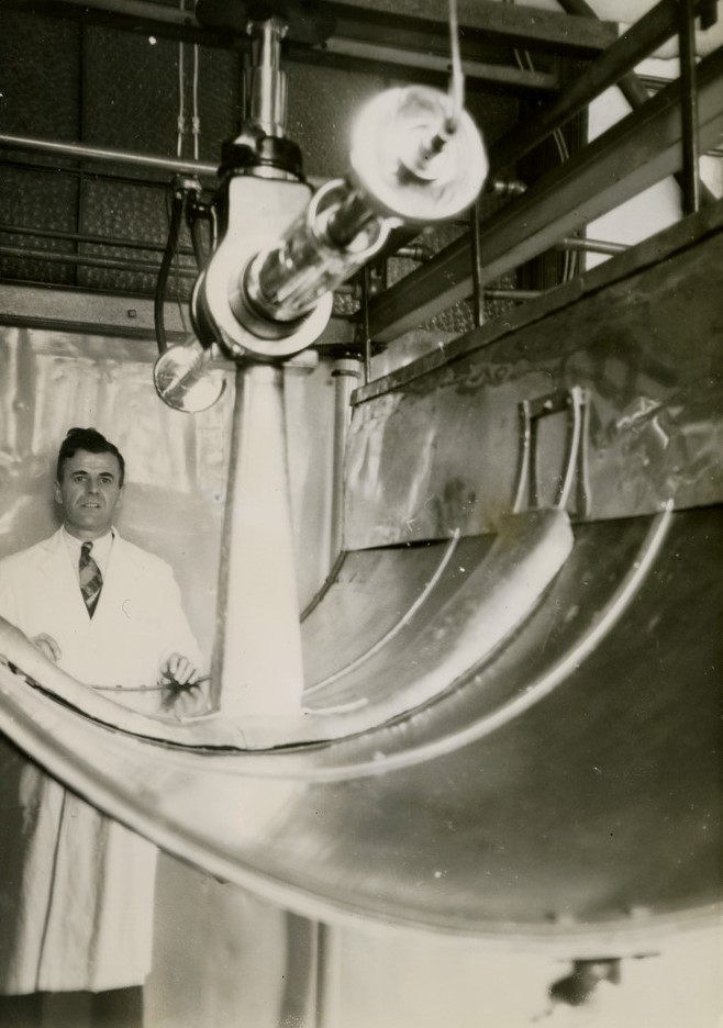 Black and white photograph of a machine in the foreground and a man in a lab coat behind it. The machine is suspended and resembles a small cylindrical cannon.