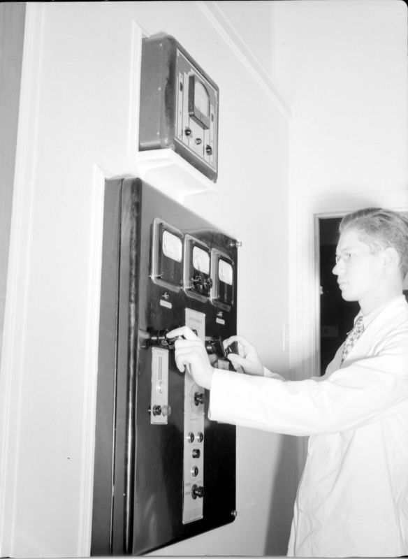 Black and white photograph of a young man standing sideways dressed in a lab coat. He is adjusting rotary knobs on a machine embedded in a white wall. The machine consists of these knobs as well as several dial gauges.