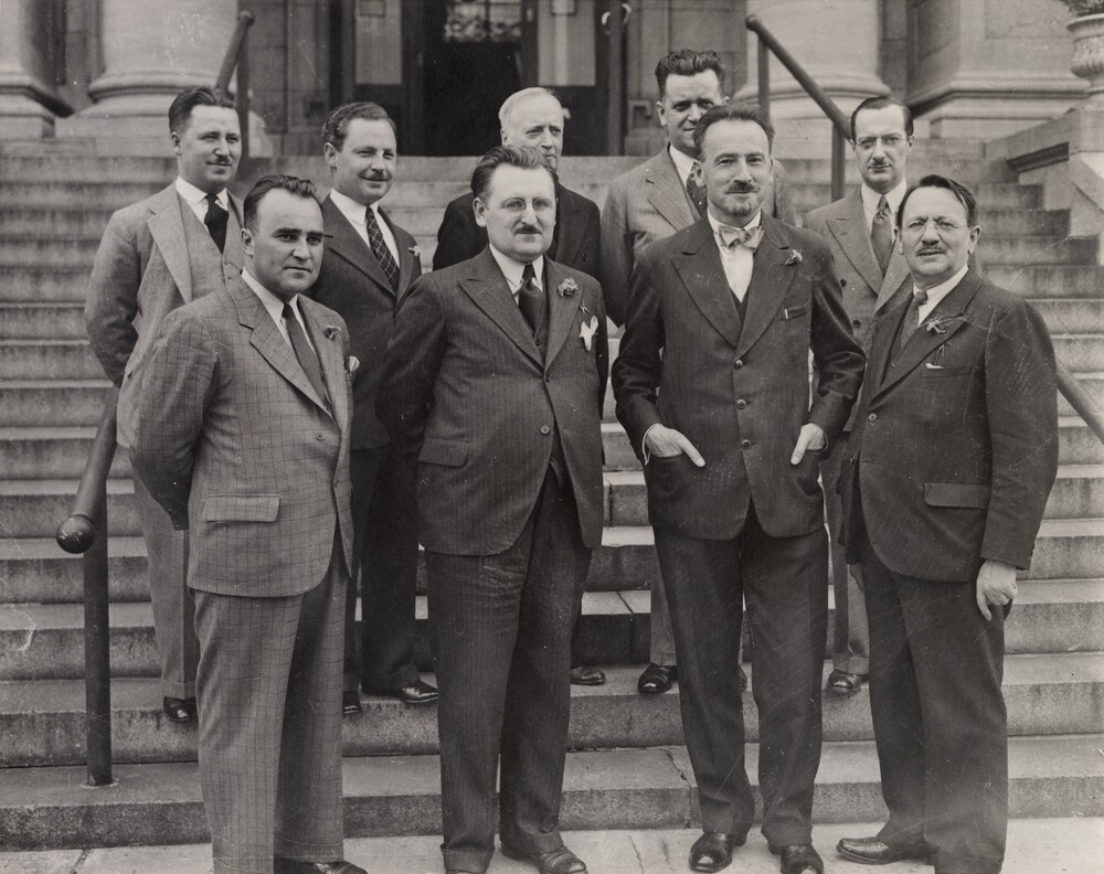 Black and white photograph depicting a group of nine men in suits standing at the bottom of a large stone staircase leading up to the door of a building, of which only the base, marked by the feet of large stone pillars, is visible.