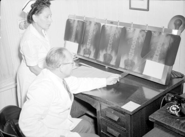 Black and white photograph depicting a woman and a man in a small room. The man, seated in a chair at a desk, is wearing a lab coat and pointing with a ruler at a light board on which transparent plastic radiographs are pinned. The woman standing is in a nurse's uniform, assisting him.