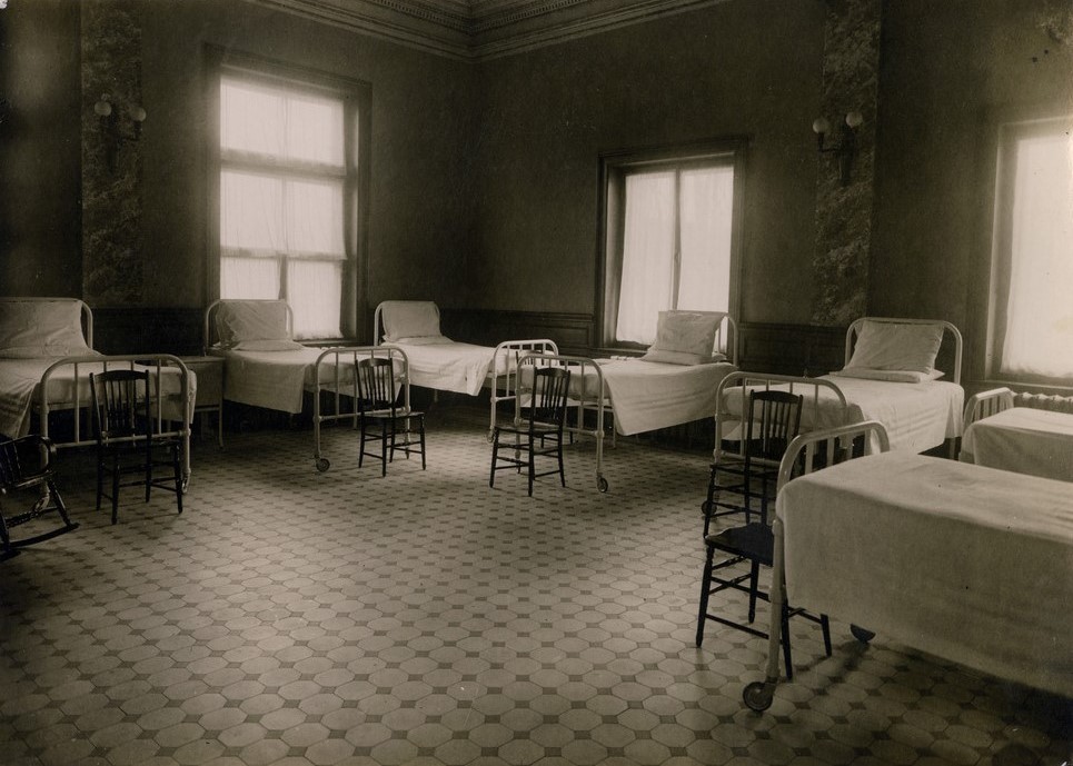 Black and white photograph depicting the interior of a room with three visible windows lining the walls. The room is very plain, furnished only with seven hospital beds and a small chair in front of each.